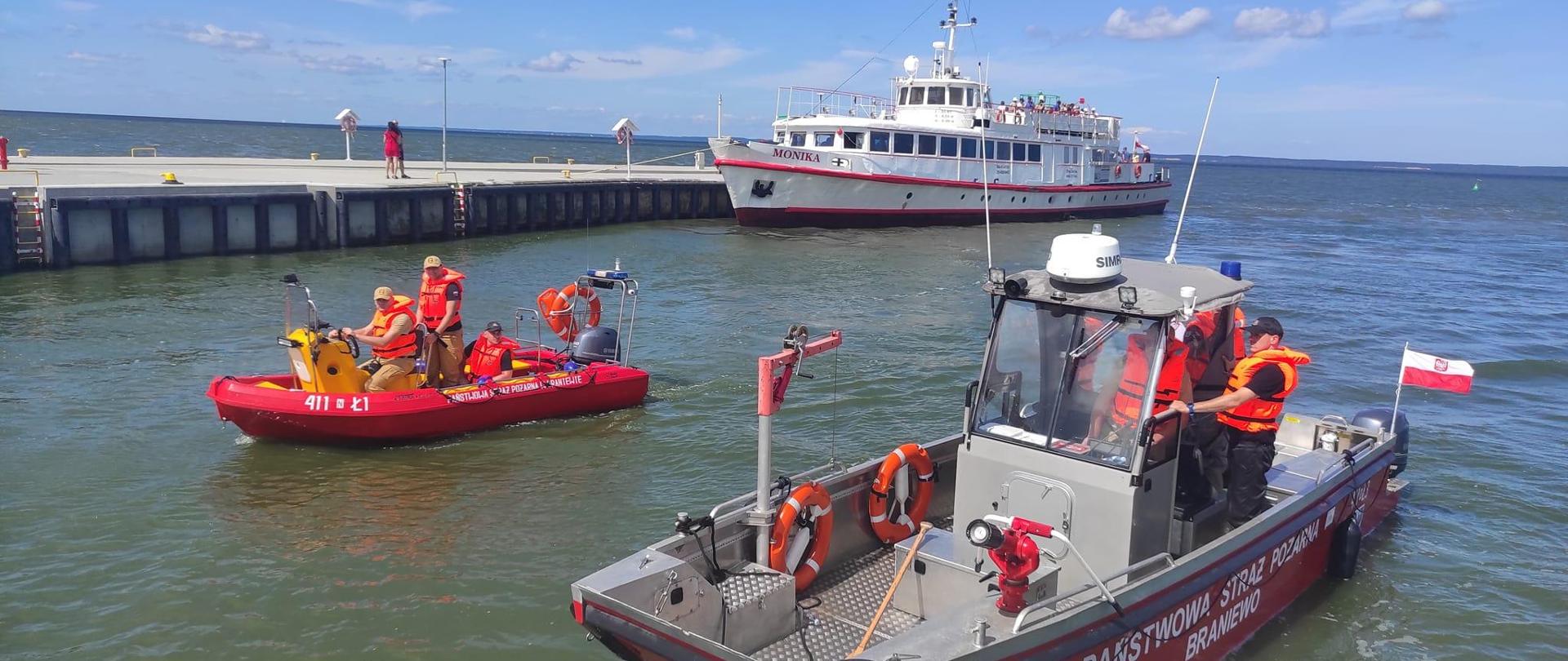 W kanale portowym stoją dwie łodzie strażackie, za nimi przycumowany biały statek pasażerski. Strażacy w kamizelkach w łodziach. W oddali horyzont, niebieskie niebo.