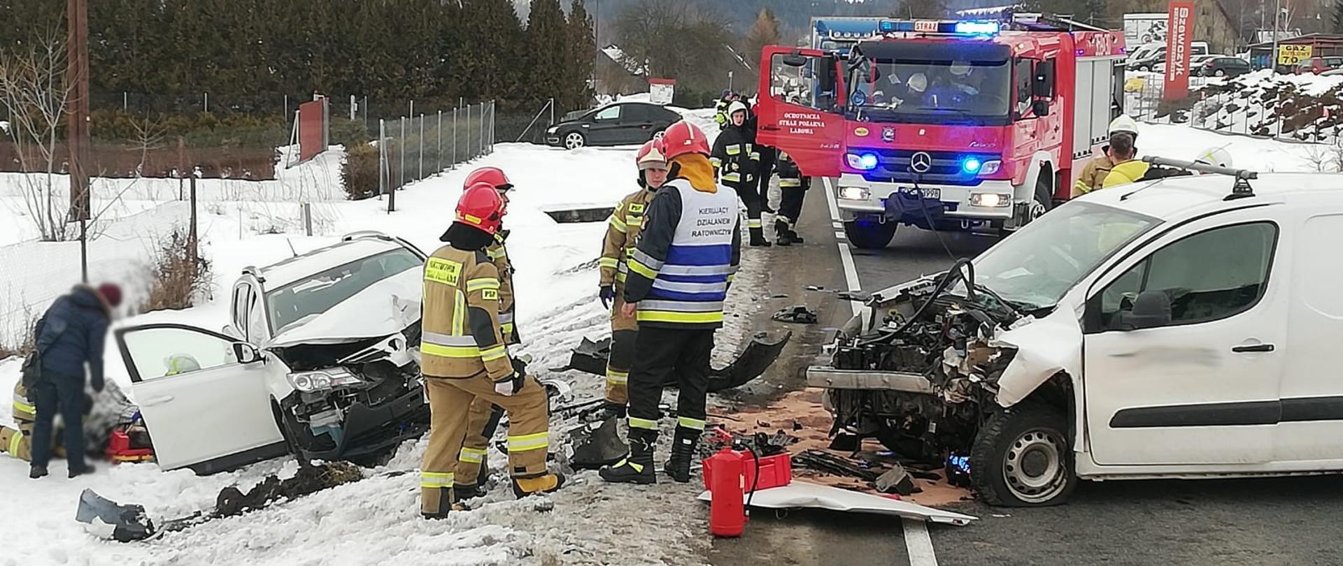 Rozbite samochody osobowe. Jeden na drodze a drugi poza drogą. Obok strażacy a w tle samochód pożarniczy.