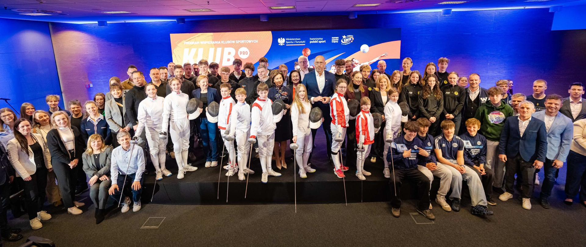 Zdjęcie zbiorowe uczestników konferencji poświęconej trzeciej edycji programu KLUB PRO. Na pierwszym planie młodzi sportowcy - szermierze. Na środku minister sportu i turystyki Jakub Rutnicki. Z tyłu grafika poświęcona programowi.