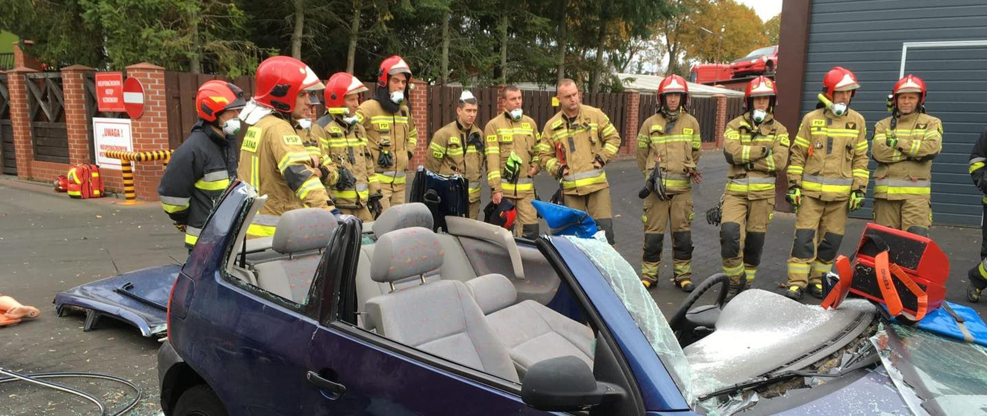 Na zdjęciu strażacy PSP po wykonanych ćwiczeniach. Na pierwszym tle widoczny samochód z wyciętym dachem oraz szybami. W tle drzewa