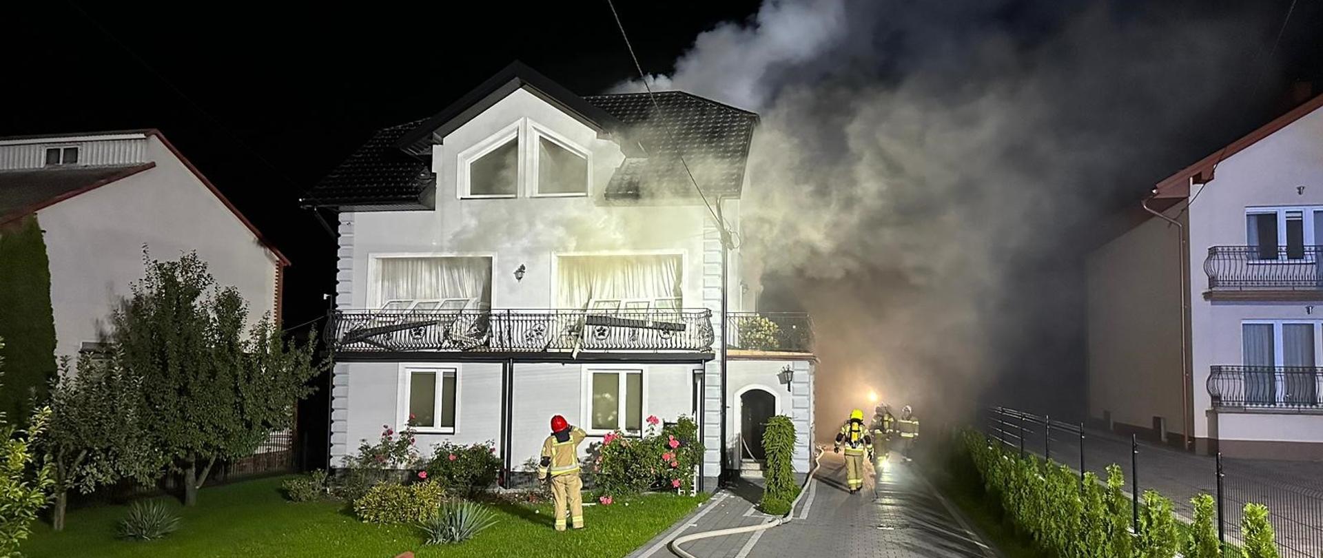 Na zdjęciu budynek jednorodzinny, z którego wydobywa się gęsty dym przez otwory okienne. Wokół domu strażacy w ubraniach specjalnych oraz aparatach powietrznych usuwający skutki pożaru. Zdjęcie zrobione jest w porze nocnej. 
