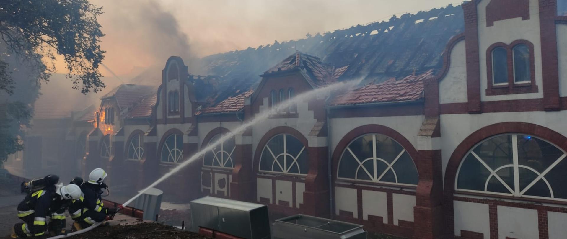 Zdjęcie przedstawia dwóch strażaków gaszących z dachu garażu budynek basenu