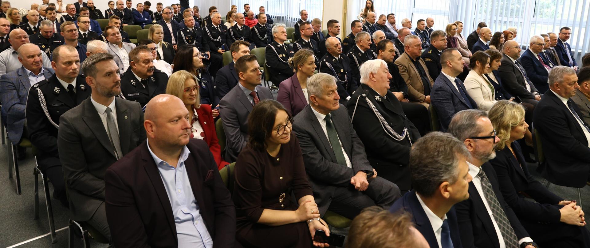 Sala konferencyjna wypełniona uczestnikami uroczystości. W pierwszych rzędach siedzą przedstawiciele instytucji, samorządów i organizatorzy wydarzenia, za nimi druhny i druhowie OSP w galowych mundurach. Wszyscy zwróceni są w stronę mównicy.