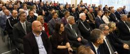 Sala konferencyjna wypełniona uczestnikami uroczystości. W pierwszych rzędach siedzą przedstawiciele instytucji, samorządów i organizatorzy wydarzenia, za nimi druhny i druhowie OSP w galowych mundurach. Wszyscy zwróceni są w stronę mównicy.