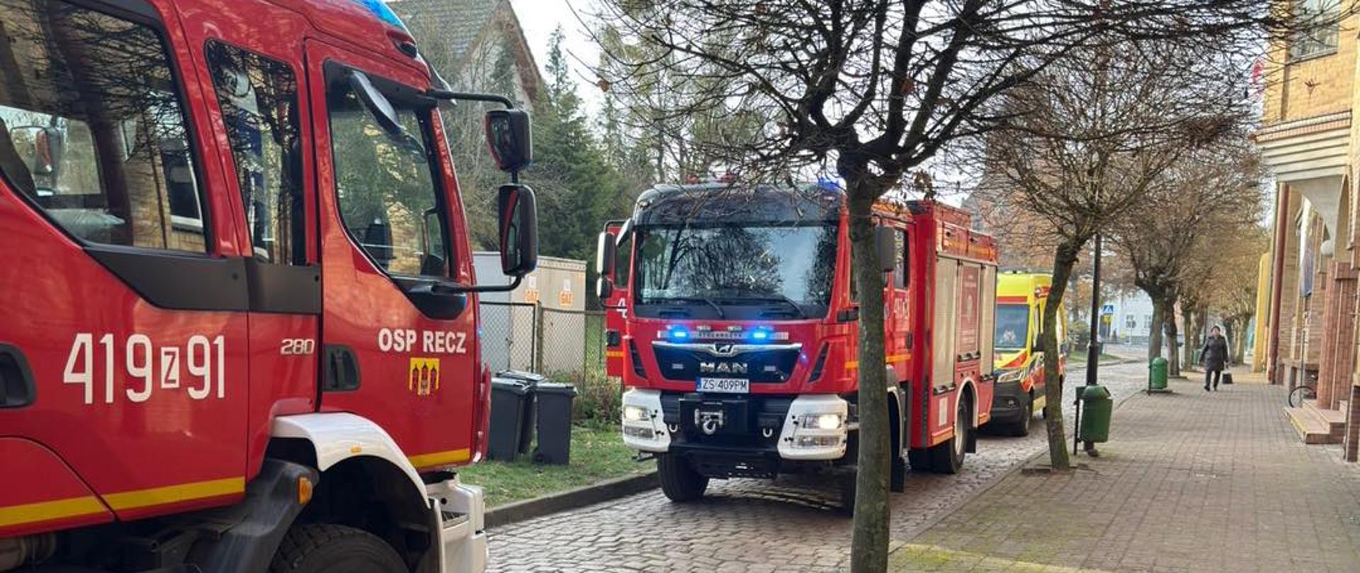 Dwa pojazdy gaśnicze oraz karetka pogotowia stoją na drodze przed budynkiem mieszkalnym.
