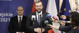 Minister funduszy i polityki regionalnej Grzegorz Puda na konferencji inaugurującej Fundusze Europejskie dla Podlaskiego 2021-2027, minister stoi przed mikrofonami z dziennikarzami, za nim stoi marszałek województwa podlaskiego Artur Kosicki