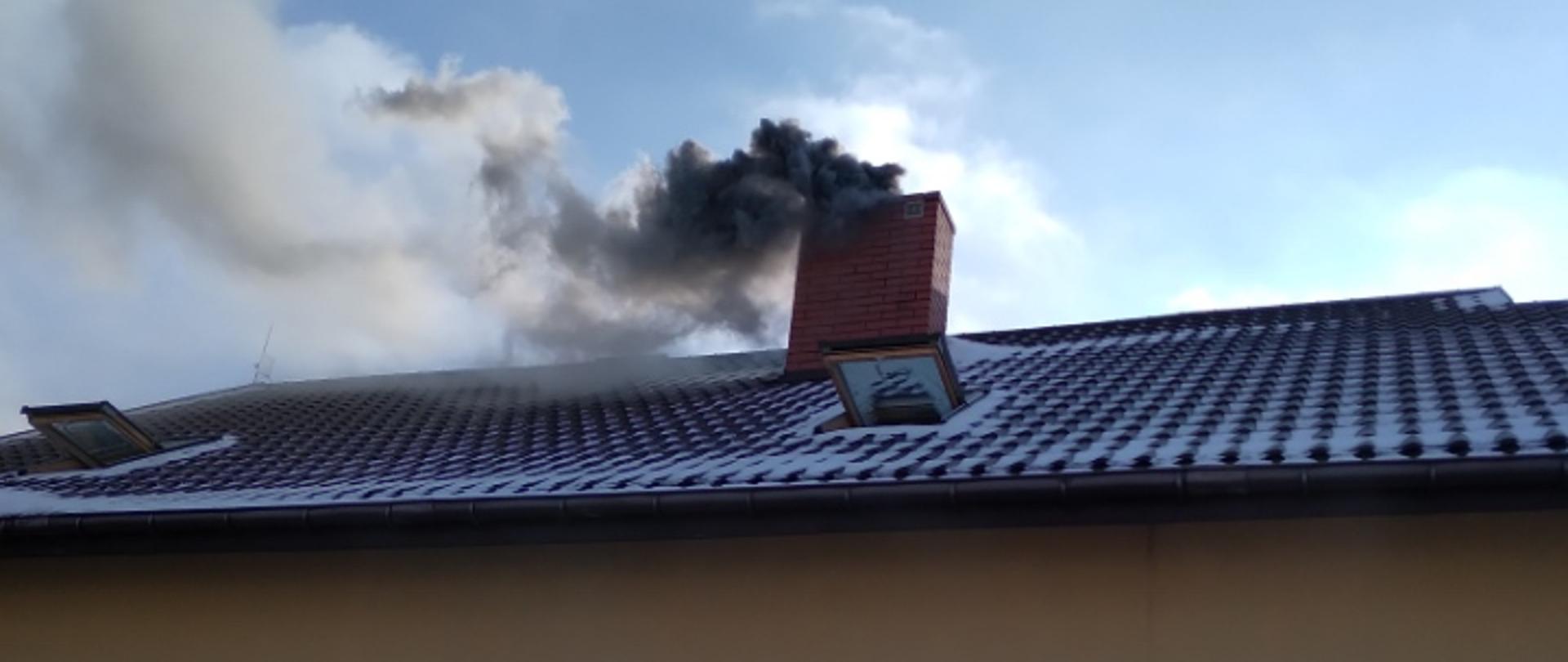 Zdjęcie przedstawia szary, gęsty dym wydobywający się z komina. Przed budynkiem w którym doszło do pożaru stoi dwóch strażaków.