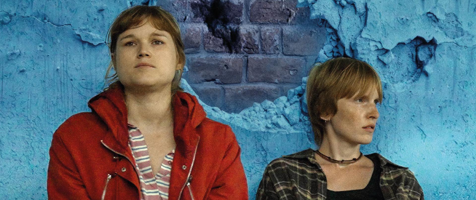 Plakat filmu „Dos hermanas” pokazuje dwie młode kobiety siedzące na ziemi na tle niebieskiej, odrapanej ściany z dużym ubytkiem tynku, przez który widać cegły. Kobieta po lewej ma na sobie czerwoną kurtkę i patrzy przed siebie z poważnym, zamyślonym wyrazem twarzy, a kobieta po prawej ma krótkie jasne włosy, ciemną koszulę w kratę i spogląda w bok. Nad nimi, dużymi żółtymi literami, widnieje tytuł „DOS HERMANAS”, a wyżej informacja, że to film w reżyserii Łukasza Karwowskiego. Plakat informuje o pokazie 26 lutego o godz. 18:00 w Alianza Francesa Polanco w Meksyku oraz o bezpłatnym wstępie. Na dole znajdują się logotypy instytucji, w tym ambasad Polski i Ukrainy oraz Unii Europejskiej.