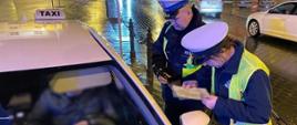 Umundurowany inspektor lubelskiej Inspekcji Transportu Drogowego i funkcjonariusz Policji stoją obok zatrzymanej do kontroli taksówki. Inspektor trzyma w ręku i sprawdza wymaganą licencję uprawniającą do zarobkowego przewozu osób.
