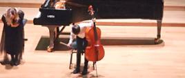 Chłopiec i kobieta stojąc przed fortepianem kłaniają się. Chłopiec trzyma wiolonczelę.