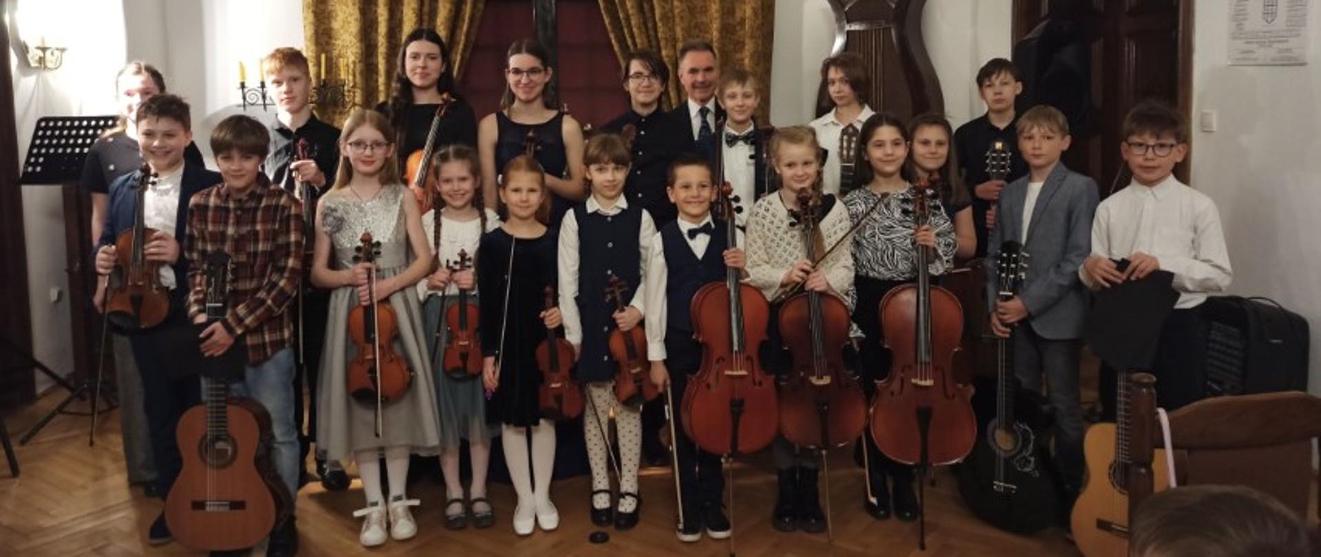 Odświętnie ubrani uczniowie z instrumentami: skrzypcami, wiolonczelami i gitarami pozują do zdjęcia. W tle wysokie okno ozdobione zasłonami.
