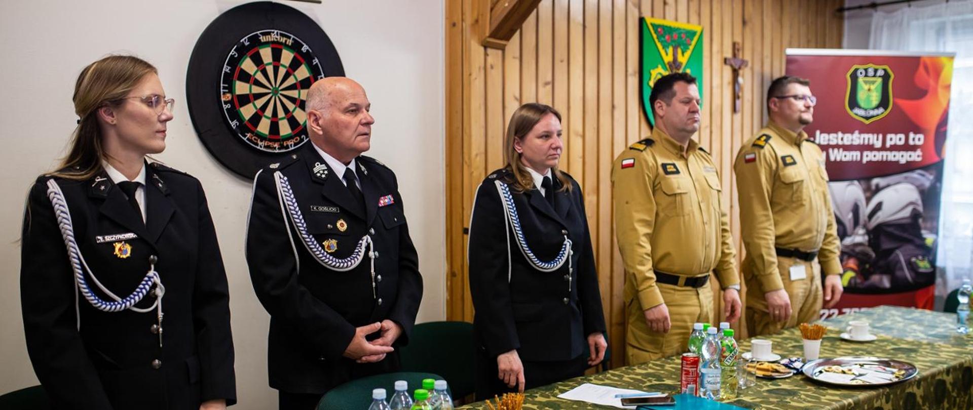 Na zdjęciu widać grupę pięciu osób stojących w pomieszczeniu o charakterze oficjalnym, prawdopodobnie podczas jakiejś uroczystości lub spotkania służbowego.
Po lewej stronie stoją trzy osoby w czarnych, galowych mundurach (najpewniej straży pożarnej). Mają przypięte odznaczenia, sznury reprezentacyjne i wyglądają bardzo formalnie. Wśród nich jest jedna starsza osoba (mężczyzna) oraz dwie kobiety. Wszyscy stoją na baczność, patrząc przed siebie.
Po prawej stronie znajdują się dwaj mężczyźni w jasnobrązowych (piaskowych) mundurach – mogą to być przedstawiciele innej służby, np. leśnej lub ratowniczej. Oni również stoją w podobnej, oficjalnej postawie.
W tle po lewej stronie wisi tarcza do rzutek (dart), a ściana częściowo jest wykończona drewnem. Po prawej stronie widać stojący baner z emblematem oraz napisem w języku polskim („Jesteśmy po to, by Wam pomagać”), co sugeruje charakter służb pomocowych.
Na stole przed nimi znajdują się różne przedmioty: butelki z wodą, kubki, przekąski oraz talerz – wygląda to jak przygotowanie do spotkania lub poczęstunku.
Cała scena sprawia wrażenie oficjalnego zebrania, apelu lub małej uroczystości w lokalnej jednostce lub instytucji.