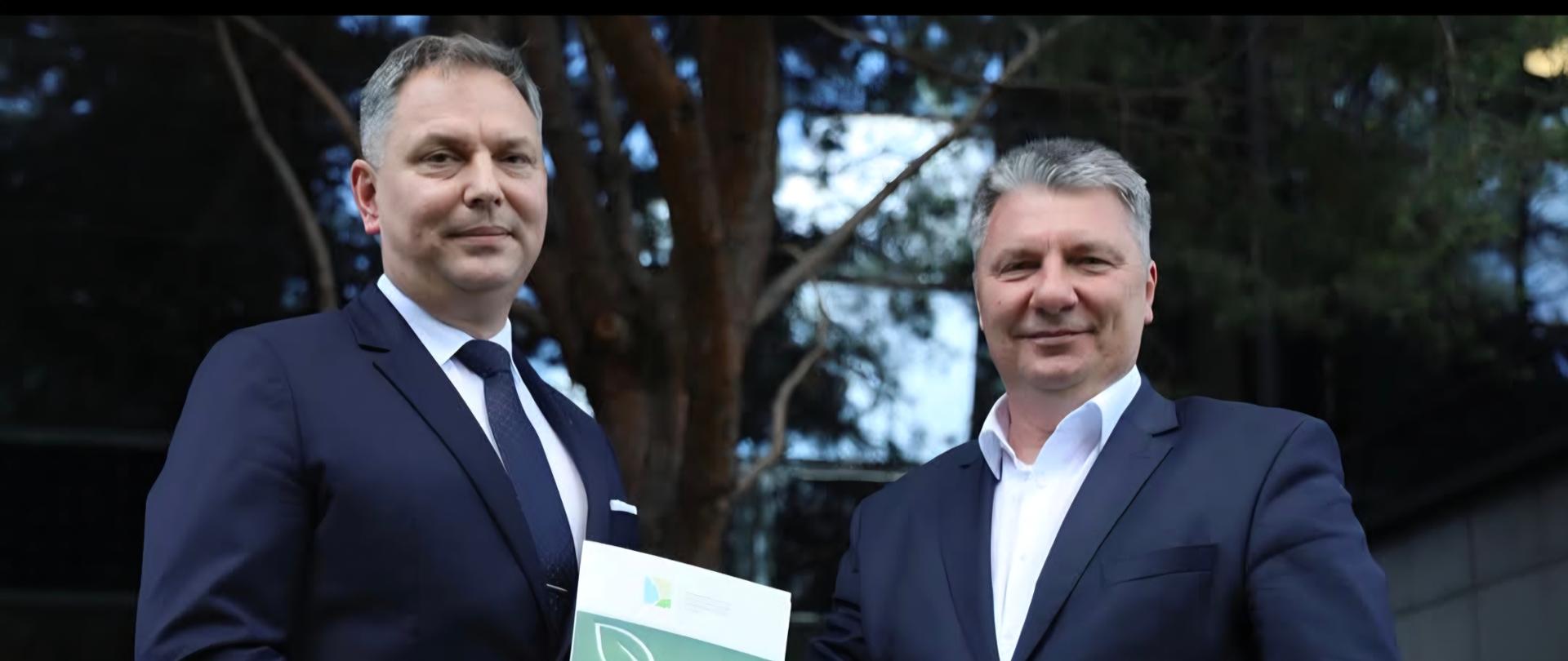 Dwóch mężczyzn w ciemnych garniturach stoi na zewnątrz na tle drzew. Mężczyzna po lewej trzyma zielono-białą publikację z graficznym wzorem, a obaj patrzą w stronę aparatu.