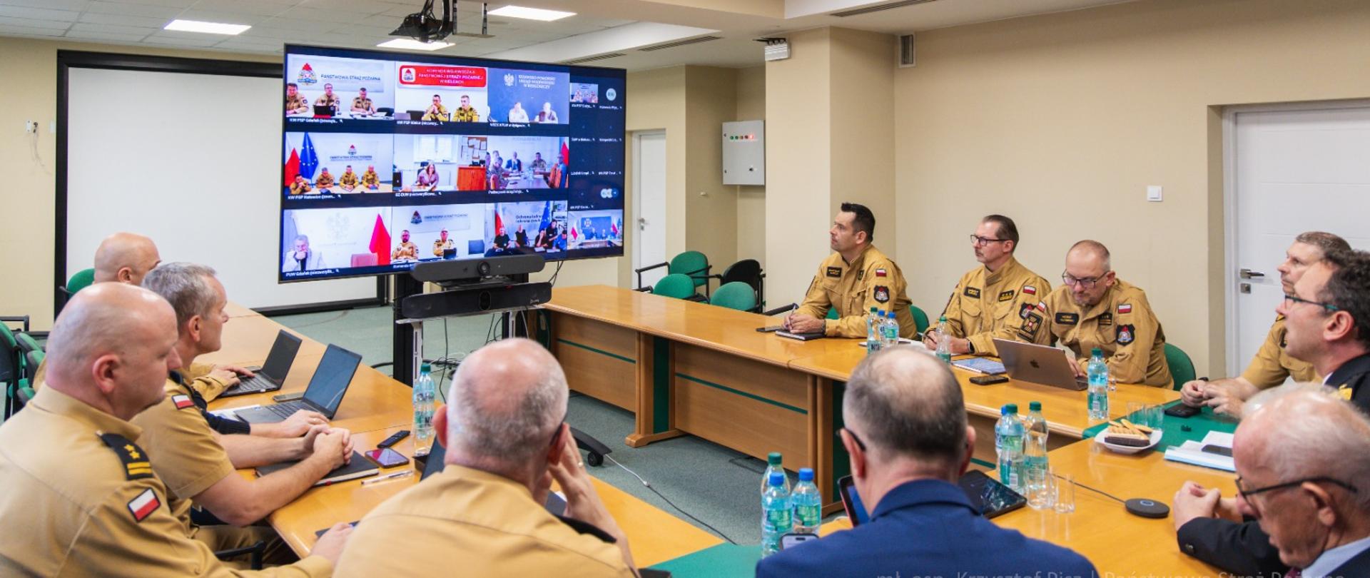 Uczestnicy spotkania siedzą przy stołach w sali konferencyjnej i obserwują duży ekran, na którym widoczni są uczestnicy wideokonferencji. 