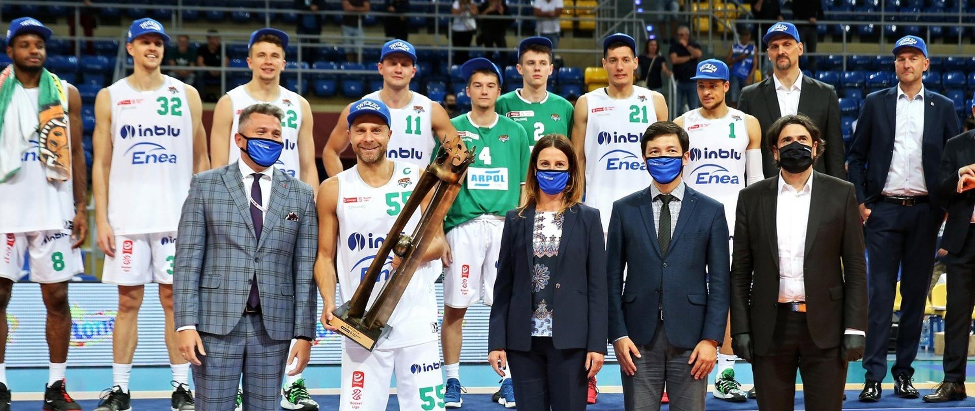 Pani Minister Sportu Danuta Dmowska Andrzejuk stoi na parkiecie boiska wraz z zawodnikami. Zawodnik z nr 55 trzyma w ręku puchar. 