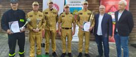 Zdjęcie przedstawia strażaków oraz osoby cywilne podczas wręczenia pucharów i dyplomów za zajęcie pierwszego miejsca. 