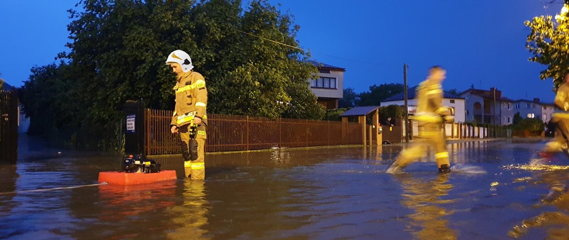 Strażak w ubraniu specjalnym hełmie przy motopompie wypompowującej wodę z zalanej ulicy i posesji, obok drugi ratownik przemieszczający się przez rozlewisko