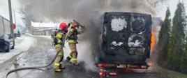 Na auto lawecie stoi płonący samochód typu bus przy nim dwóch strażaków gasi płonący samochód z boku widoczne płomienie oraz drzewka nad samochodem kłęby dymu