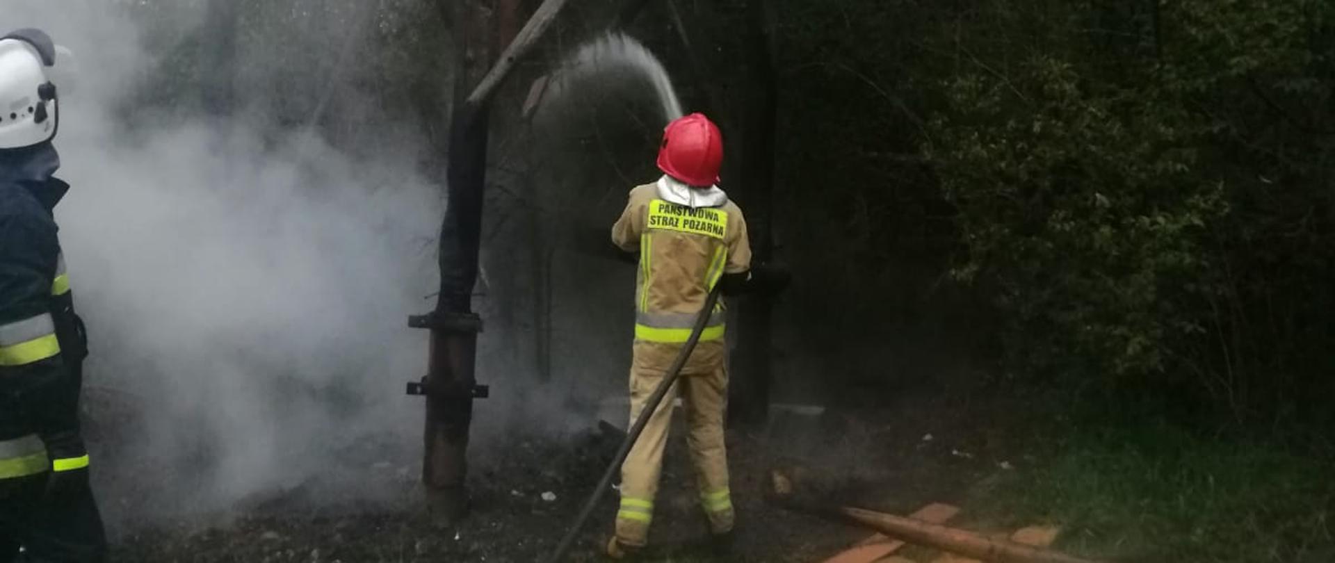 Kolorowe zdjęcie przedstawia widok spalonej ambony myśliwskiej na tle zielonych drzew. Strażacy gaszą wodą palące się resztki konstrukcji nośnej ambony myśliwskiej. Widoczne spalone częściowo drewniane słupy i pogorzelisko. Obok ambony leżą rozrzucone deski.