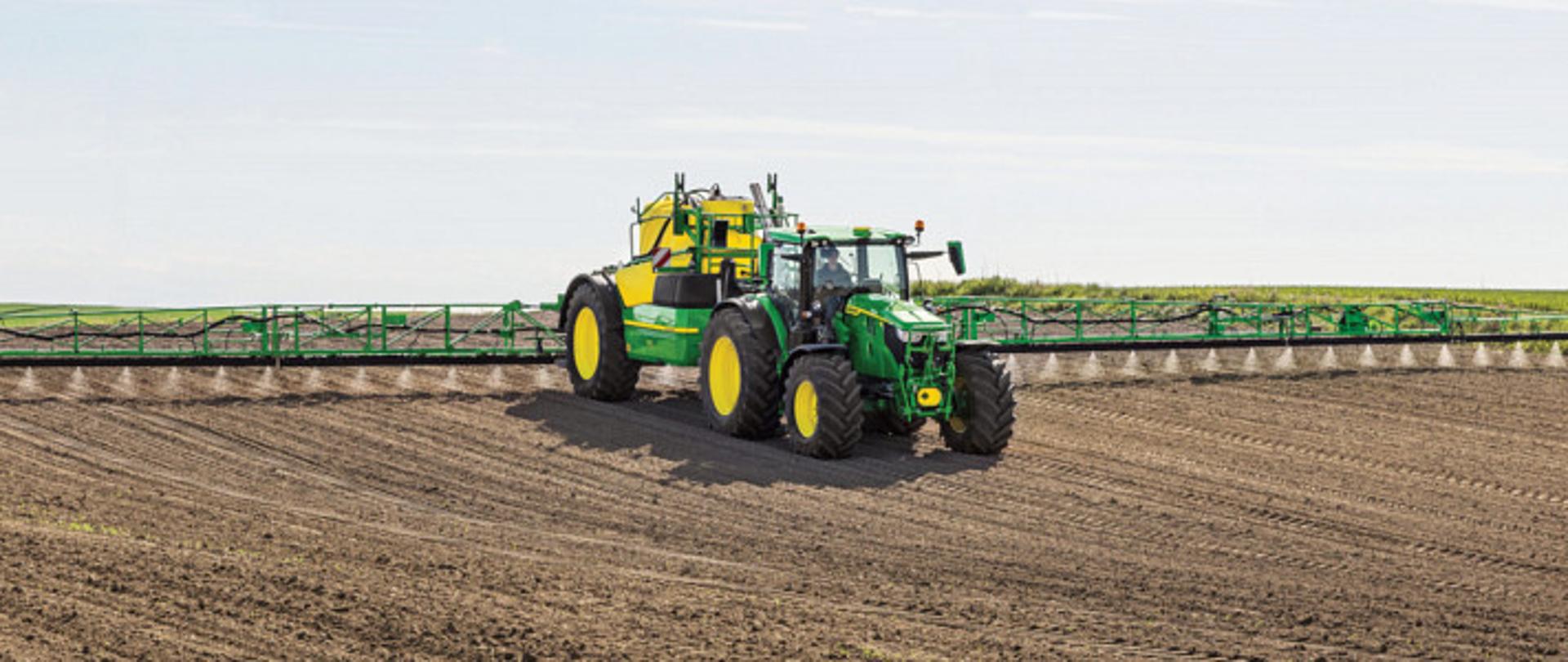 Na zdjęciu znajduje się ciągnik rolniczy marki John Deere z przyczepionym opryskiwaczem polowym. Maszyna porusza się po dużym, starannie uprawionym polu. Opryskiwacz jest rozłożony na całą szerokość roboczą, co pozwala na równomierne rozprowadzanie środków ochrony roślin na dużej powierzchni. W tle widoczne są pasy zieleni oraz niebo z lekkim zachmurzeniem.