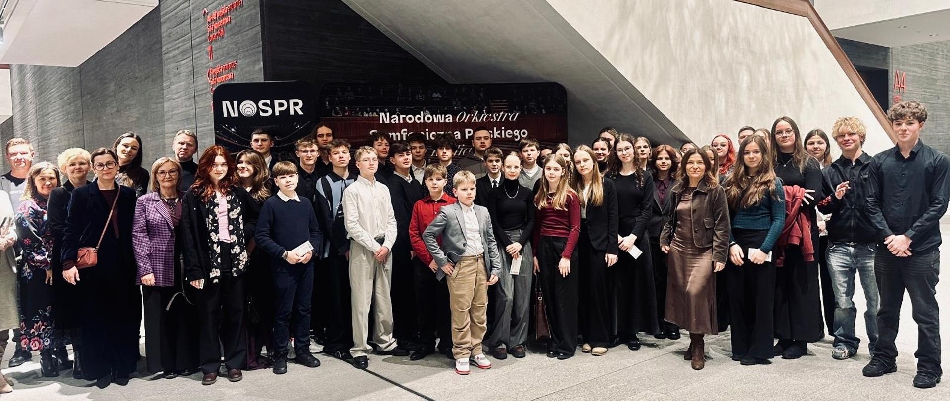 Grupa osób, w tym młodzież i dorośli, pozuje wspólnie do zdjęcia w nowoczesnym wnętrzu budynku Narodowej Orkiestry Symfonicznej Polskiego Radia (NOSPR). W tle widoczny jest baner z nazwą instytucji oraz betonowe ściany i schody o geometrycznej konstrukcji. Uczestnicy stoją w kilku rzędach, ubrani elegancko, uśmiechają się do obiektywu.