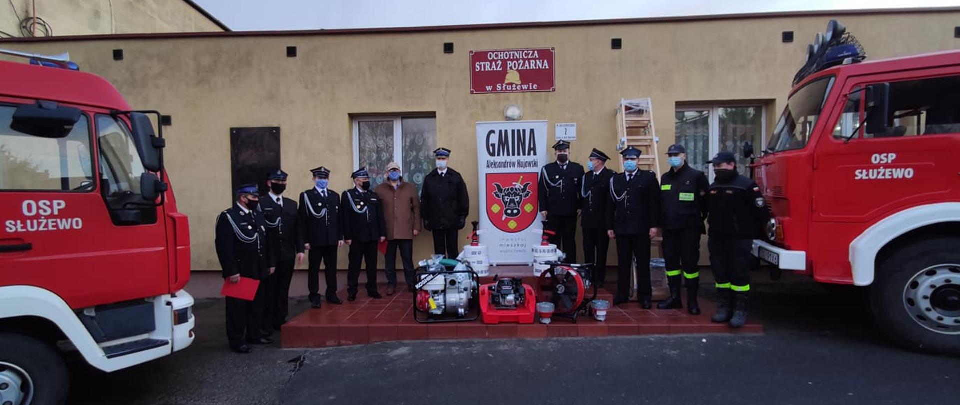 Zdjęcie w centralnej części przedstawia baner z logiem gminy Aleksandrów Kujawski. Po bokach stoją strażacy w umundurowaniu wyjściowym i kosztarowym. po środku ustawiony jest nowopozystkany sprzęt. Na skrajnych brzegach zdjęcia widoczne samochody strażackie z nazwą Jednostki umieszczoną na drzwiach.
