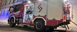 Na tle kamienicy stoi ciężki samochód ratowniczo-gaśniczy uczestniczący w akcji. Pojazd ma otwartą pierwszą skrytkę na sprzęt oraz włączone sygnały błyskowe. Od samochodu jest rozwinięta linia wężowa. Pora nocna.