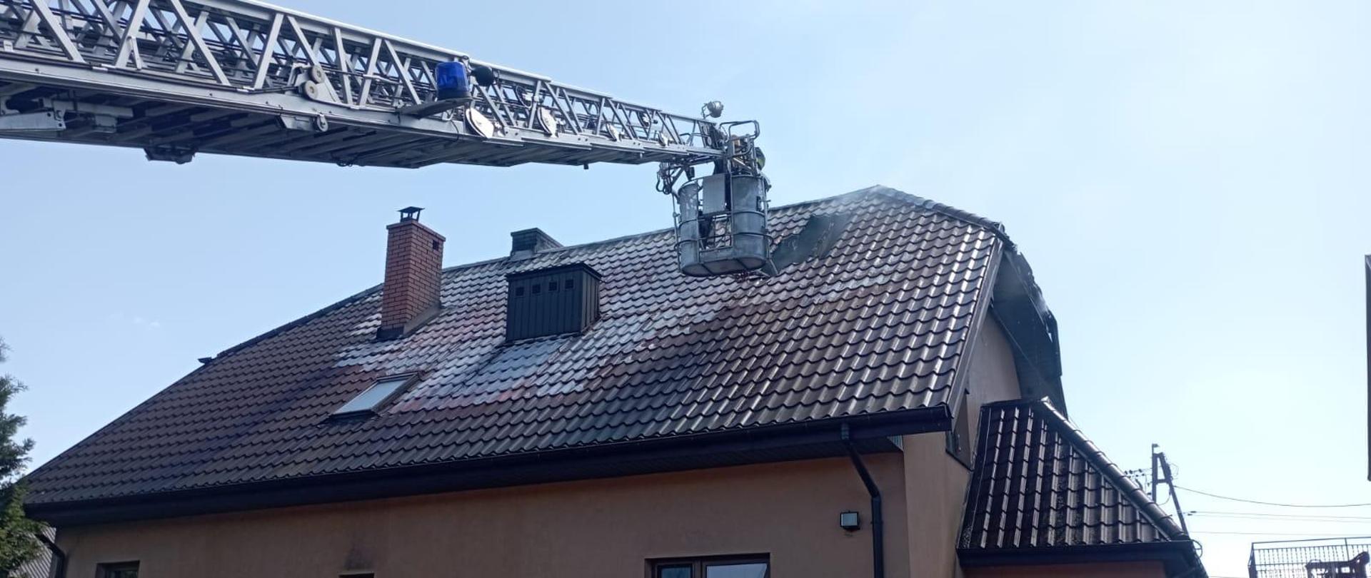 Zdjęcie przedstawia budynek mieszkalny, w którym wybuchł pożar. Zniszczenia widać na pokryciu dachowym . Fragment pokrycia został zdemontowany. Rozstawiona została drabina mechaniczna, kosz zwisa nad otworem.