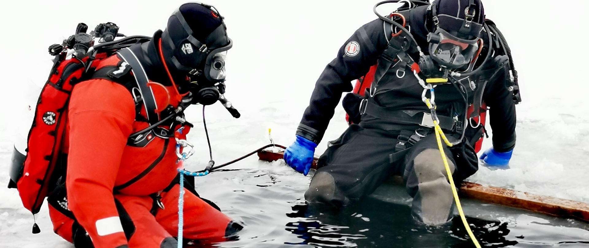 Na zdjęciu widać dwóch nurków w kombinezonach w wykutym przeręblu na zbiorniku wodnym