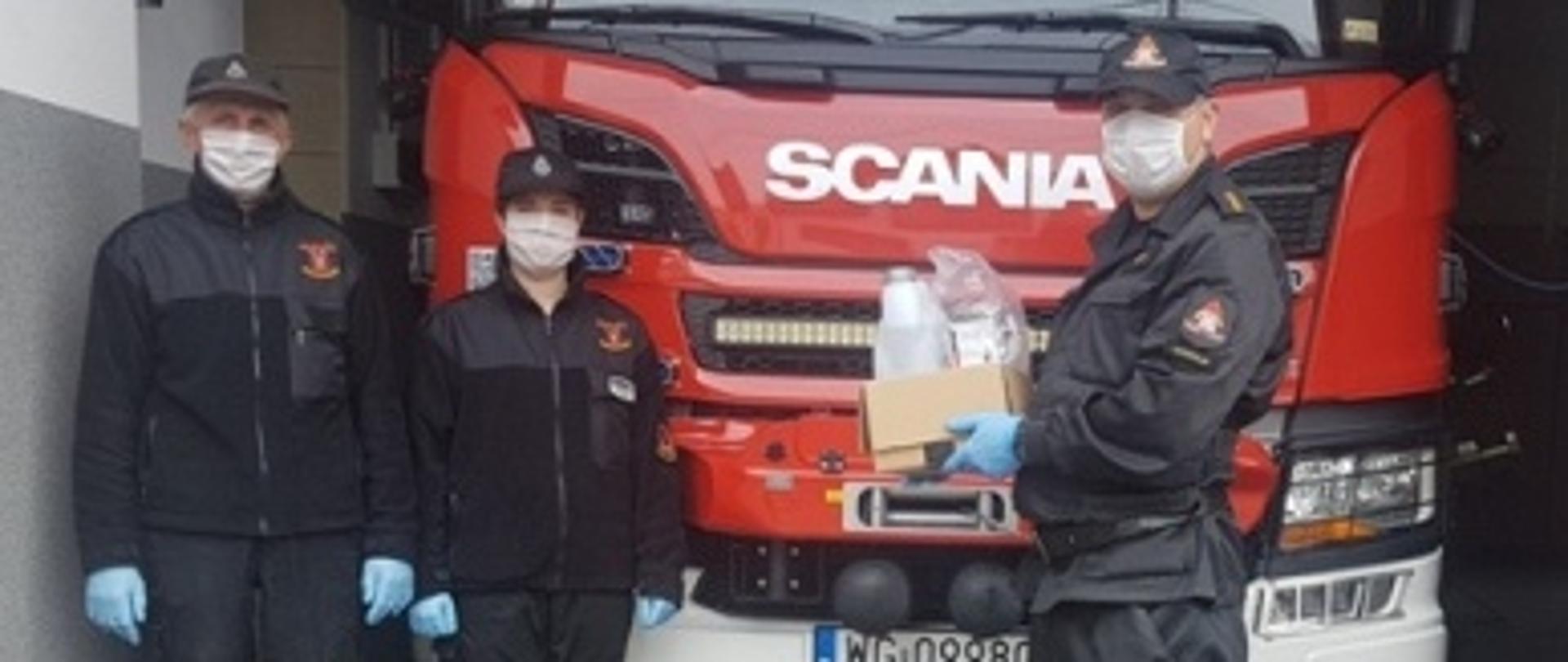 Funkcjonariusz PSP ubrany w mundur koloru czarnego przekazuje środki ochrony indywidualnej ochotnikom na tle czerwonego samochodu strażackiego