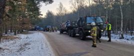 na drodze w lesie stoi samochód ciężarowy z naczepą należący do wojska przed nim stoi strażak oraz dwaj żołnierze pomiędzy samochodem z naczepą stoją strażacy zabezpieczając ładunek za nimi inne samochody wojskowe