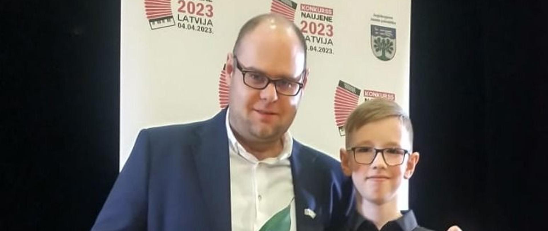 Zdjęcie kolorowe. Nauczyciel wraz z uczniem na scenie. Obaj trzymają w ręku dyplomy i kwiaty. 