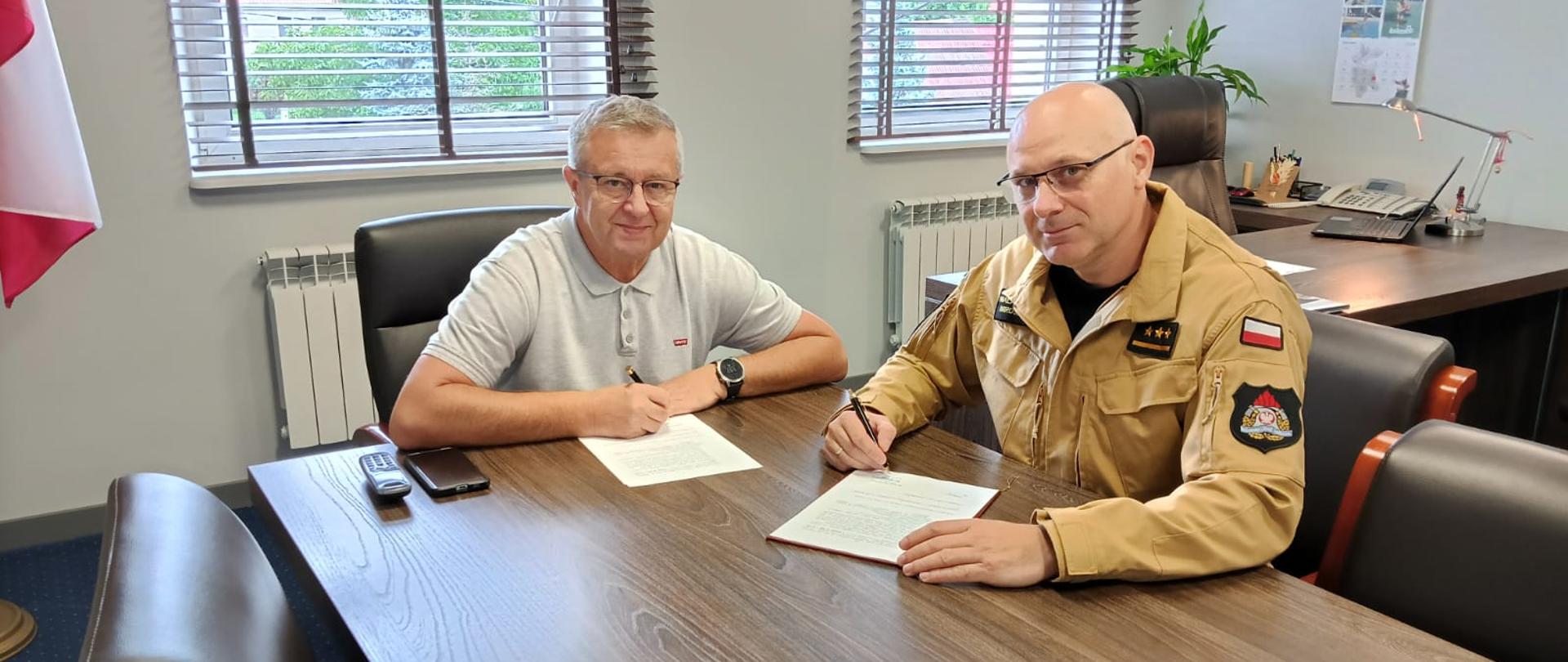 Obraz przedstawia Komendanta Powiatowego i Wójta Gminy Męcinka w trakcie podpisania porozumienia. Osoby siedzą przy stole w pomieszczeniu biurowym.