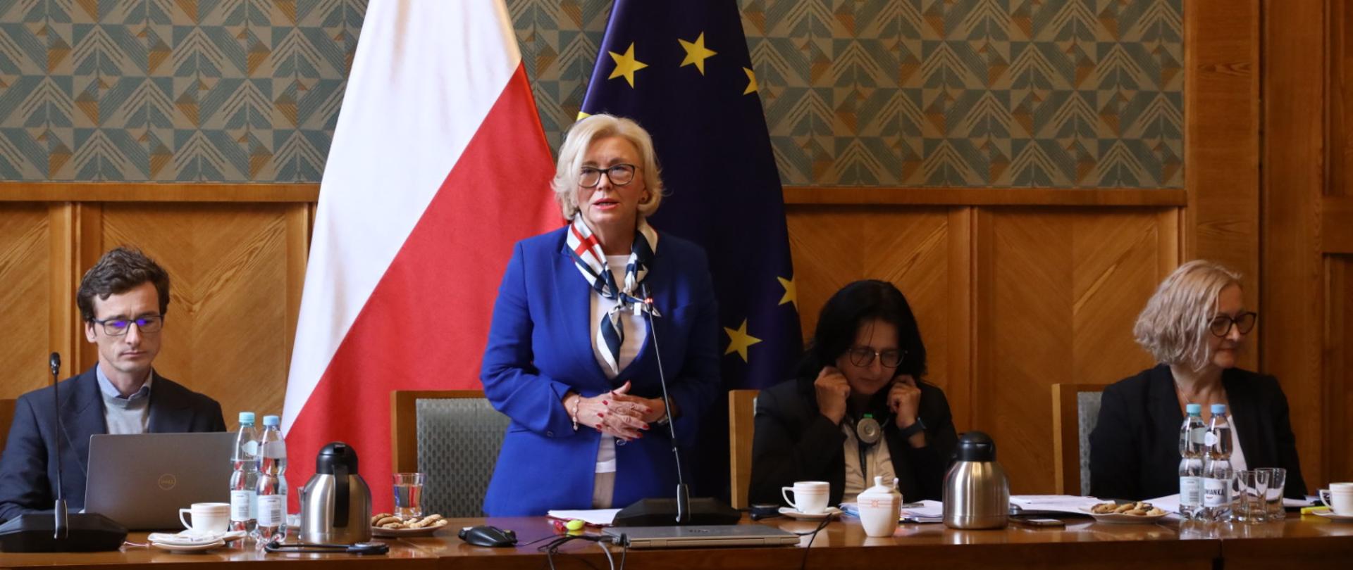 Wiceminister Machałek w niebieskim ubraniu stoi za stołem, za nią ściana w zielony wzorek i flagi Polski i UE.