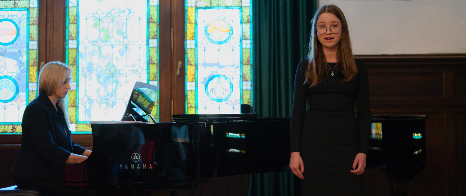 Młoda dziewczyna w okularach i czarnej sukience podczas występu wokalnego. W tle fragment fortepianu, na którym gra kobieta o blond włosach, okno z witrażem i zielone zasłony.