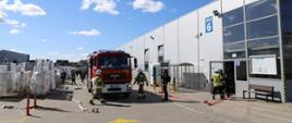 Zdjęcie przedstawia strażaków w ubraniach bojowych podczas ćwiczeń przy jednej z hal firmy Dobroplast. Strażacy rozwijają linie gaśnicze z samochodów ustawionych wzdłuż hali. Zdjęcie w słoneczny dzień wykonano na terenie firmy Dobroplast. 