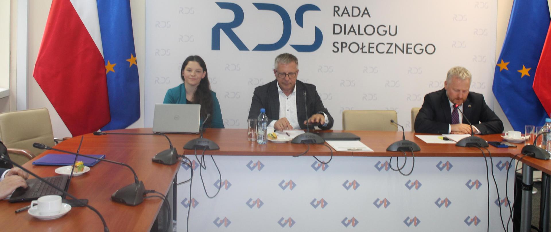 Na zdjęciu widnieje stół prezydialny za którym siedzą od lewej strony: Katarzyna Nowakowska - Podsekretarz Stanu w Ministerstwie Rodziny, Pracy i Polityki Społecznej, Przewodniczący zespołu ds. budżetu, wynagrodzeń i świadczeń socjalnych – Leszek Miętek, Jurand Drop – Podsekretarz Stanu w Ministerstwie Finansów oraz. Po obu stronach stołu widnieją dwie flagi: Polski i UE. W tle siedzących znajduje się baner z napisem Rada Dialogu Społecznego a nad nim wisi godło Polski.