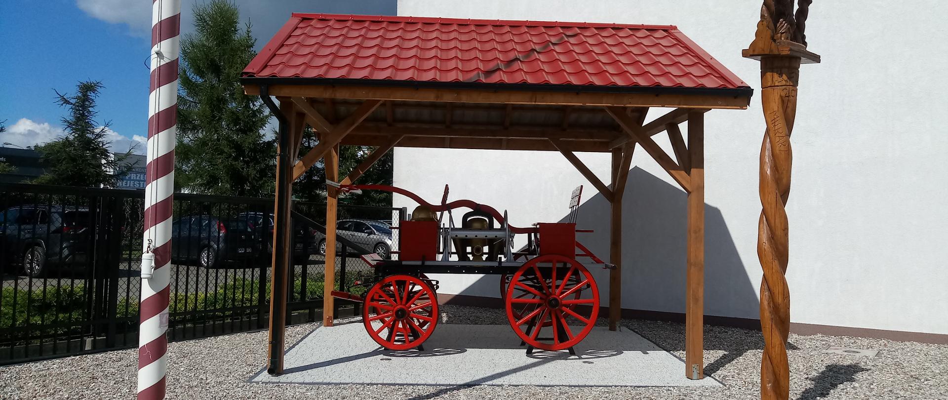 Na zdjęciu, pod specjalną wiatą, stoi stara, odrestaurowana sikawka konna,. Z boku widoczny fragment masztu flagowego i figury, stanowiącej wyposażenie placu KP PSP Inowrocław.