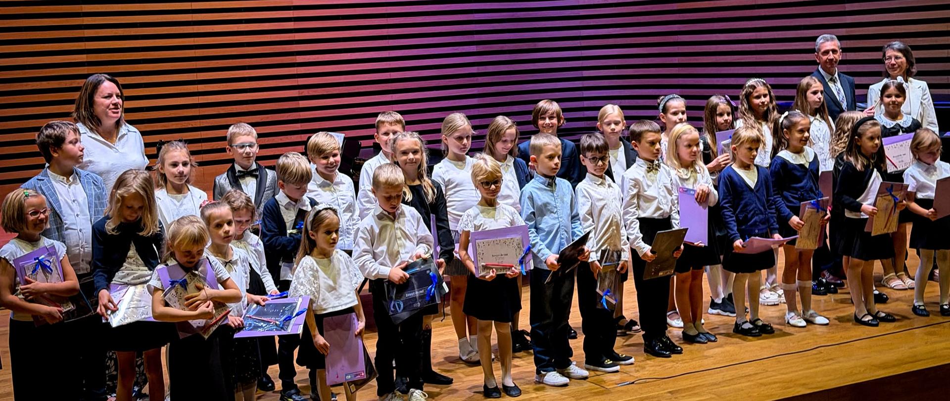 Wszyscy uczniowie stoją w dwóch rzędach z upominkami na estradzie, za nimi stoją dwie kobiety i mężczyzna.