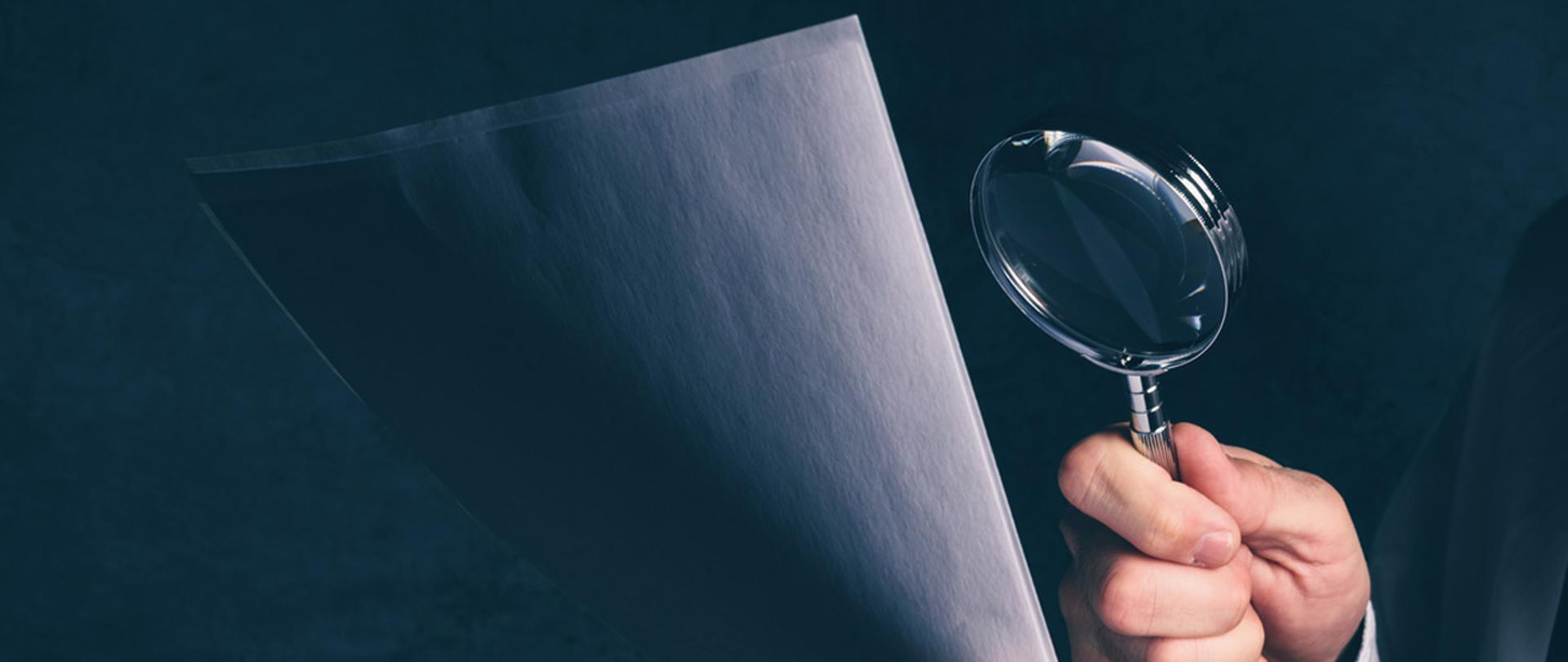 Ręka trzymająca lupę nad kartką papieru.