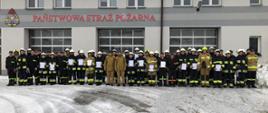 Zdjęcie przedstawia strażaków w ubraniach specjalnych na zbiórce przed garażami Jednostki Ratowniczo-Gaśniczej w Lubaczowie. 