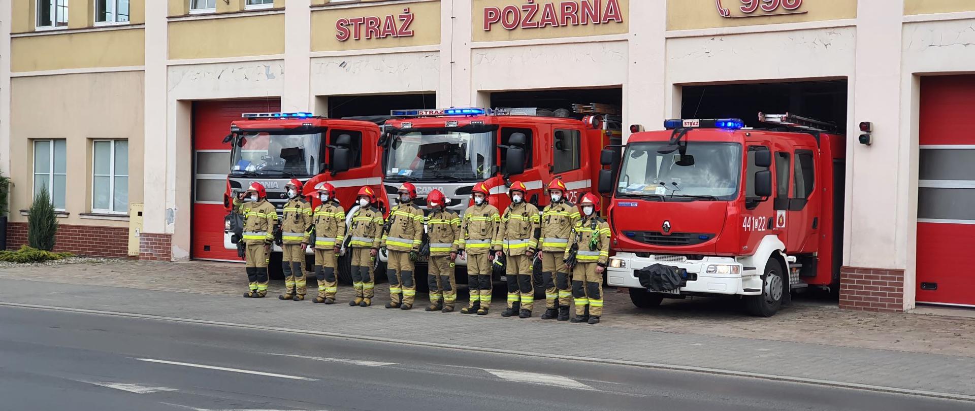 Zdjęcie przedstawia 3 czerwono- białe, ciężarowe samochody strażackie stojące w bramach wyjazdowych garażu. Auta maja włączone niebieskie światła błyskowe. Przed nimi w rzędzie stoi 10 strażaków ubranych w piaskowe mundury specjalne i czerwone hełmy i maseczki ochronne. Stojący po lewej dowódca oddaje honor.