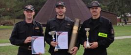 Na zdjęciu widać trzech strażaków, którzy trzymają w rękach puchary oraz dyplomy. W tle znajduje się obiekt strzelnicy oraz wigwam. 