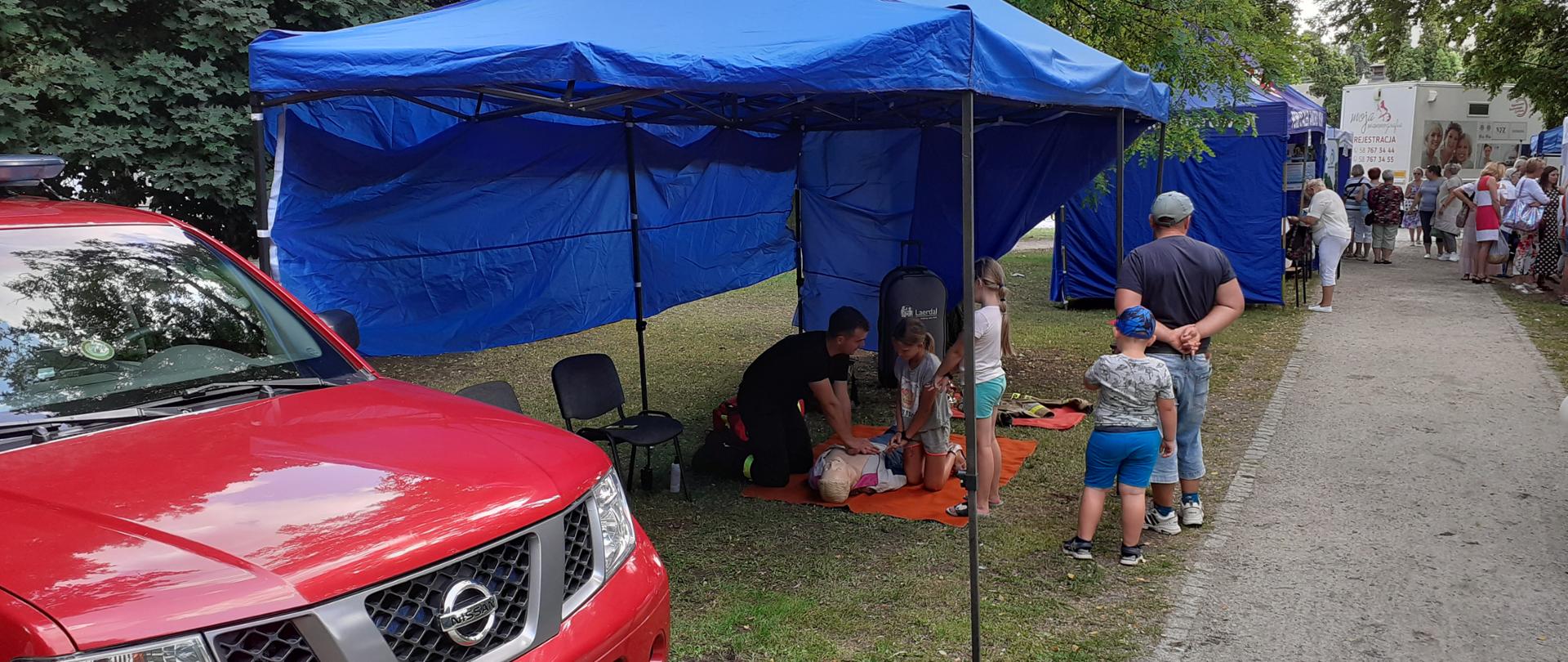 Na zdjęciu widać czerwony terenowy samochód Państwowej Straży Pożarnej oraz niebieskiego koloru namioty promocyjne ustawione wzdłuż alei parku. W namiocie na pierwszym planie widać strażaka oraz grupę osób dorosłych i dzieci. Strażak udziela instruktażu na temat udzielania pierwszej pomocy. Osoby wykonują resuscytację krążeniowo-oddechową na fantomie osoby dorosłej.