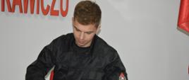 Świetlica komendy powiatowej. Przy stoliku siedzi jeden z nowo slubujących strażaków, który składa podpis na formularzu aktu ślubowania. Ma na sobie ubranie koszarowe w kolorze czarnym. 
