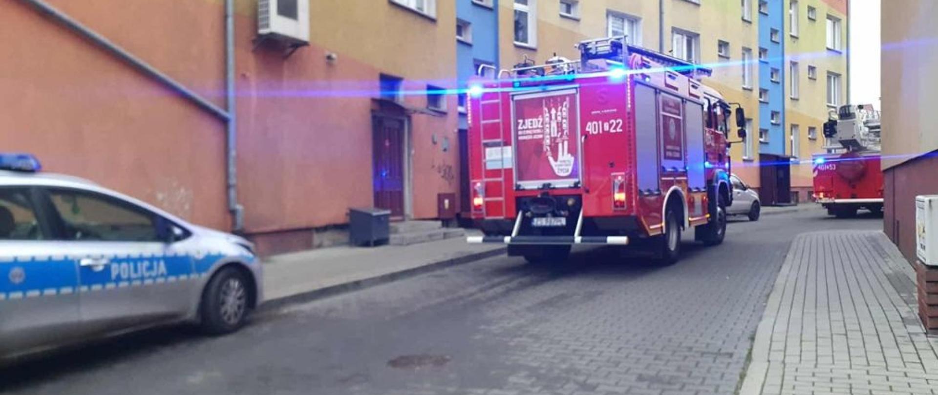 Zdjęcie przedstawia pojazdy pożarnicze przed blokiem. Od lewej strony zdjęcia widać auto policyjne. Pojazdy na sygnałach. Trwa akcja gaśnicza.