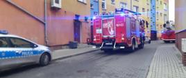 Zdjęcie przedstawia pojazdy pożarnicze przed blokiem. Od lewej strony zdjęcia widać auto policyjne. Pojazdy na sygnałach. Trwa akcja gaśnicza.