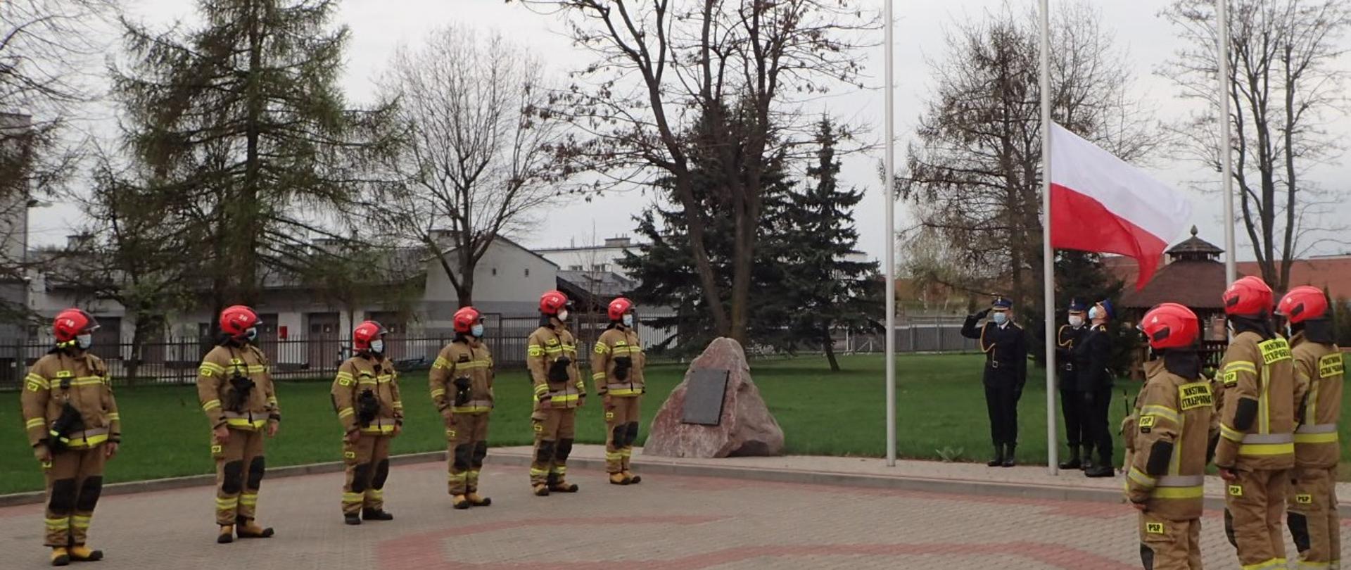 Strażacy PSP w ubraniach specjalnych stoją w szeregach naprzeciw siebie. W tle trzy maszty flagowe. Na środkowy maszt wciągana jest flaga przez poczet flagowy w mundurach wyjściowych.