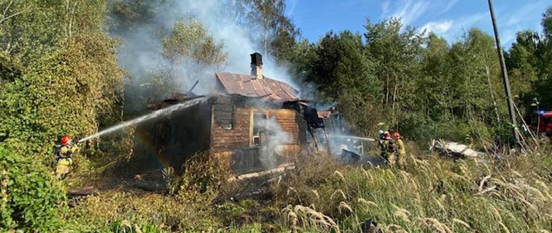 Na zdjęciu widoczny jest budynek mieszkalny jednorodzinny, konstrukcji drewnianej, który objęty był pożarem. Budynek z częściowo zniszczoną konstrukcją dachu. Na zdjęciu widoczni strażacy podający dwa prądy wody na palący się obiekt
