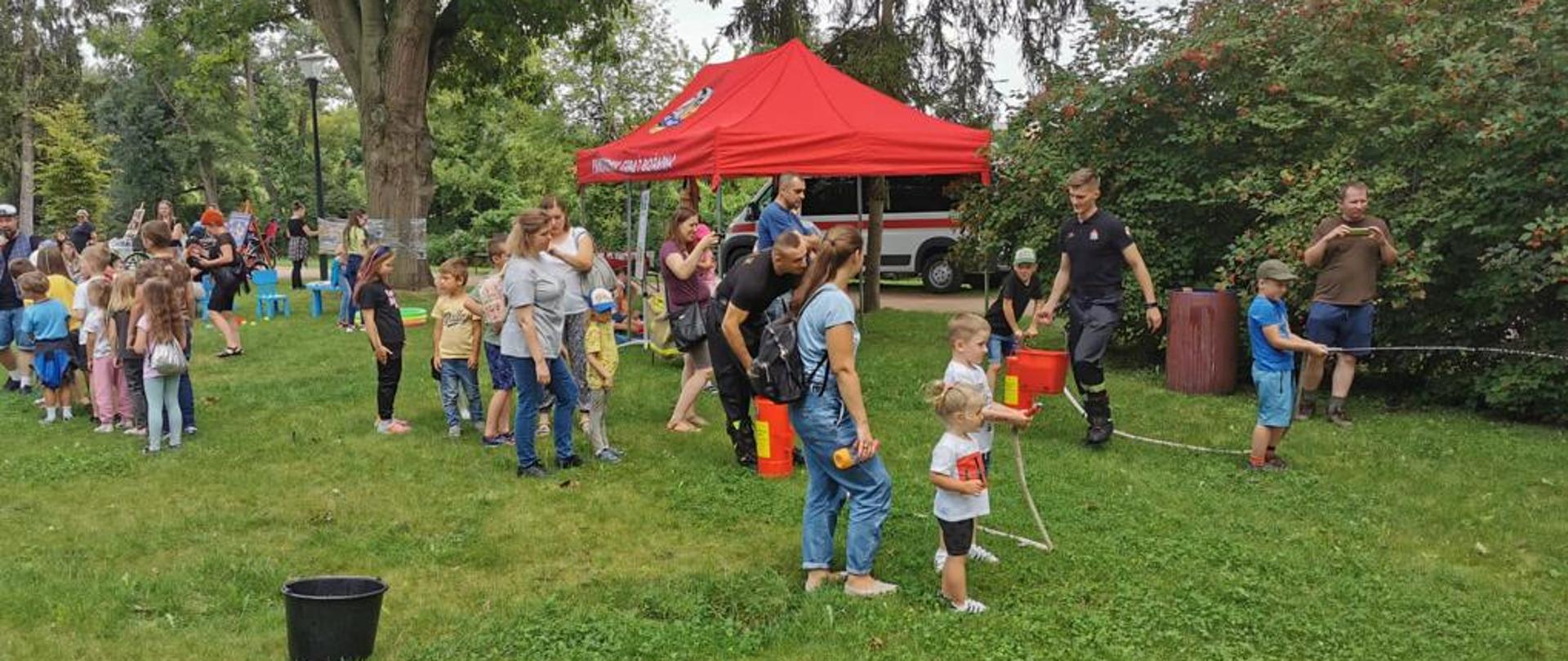 Zdjęcie przedstawia dwójkę dzieci (dwóch chłopców) trzymających małe węże z wodą i celujące w tablicę. Dookoła widać samochód strażacki oraz namioty - stoisko strażackie, strażaków dzieci i dorosłych ludzi uczestniczących w pikniku, który odbywa się w parku.